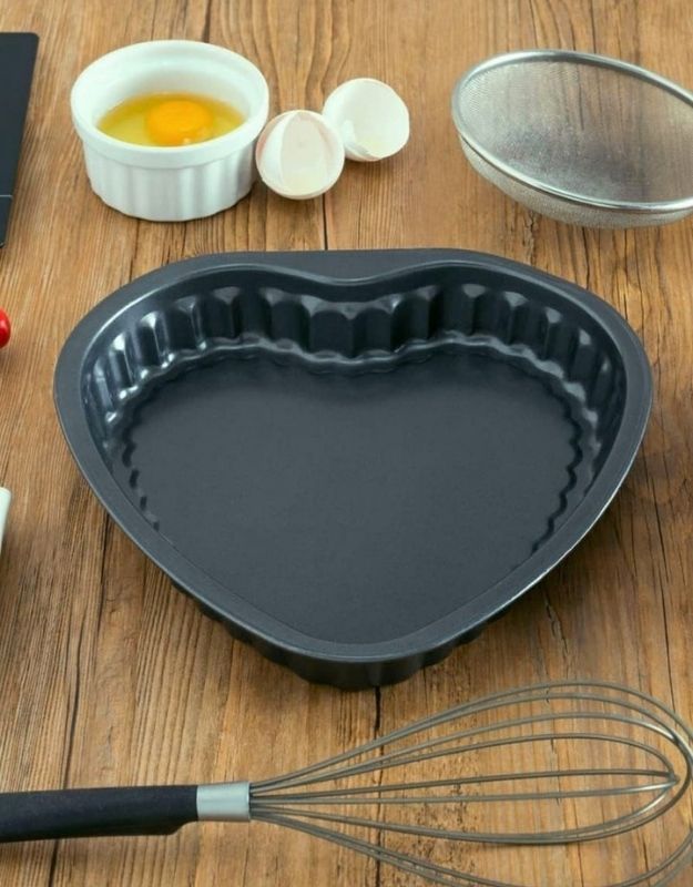 Heart-shaped baking pan on a wooden surface with eggs, a whisk, and a strainer. Nonstick heart-shaped cake pan mold on wooden table with whisk and eggs, perfect for baking cakes and desserts.